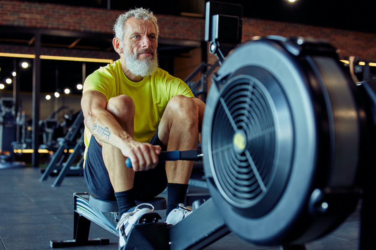 Sukan dan kecergasan selepas 50. Lelaki tua atletik matang yang kuat dalam pakaian sukan bersenam pada mesin mendayung di gimnasium